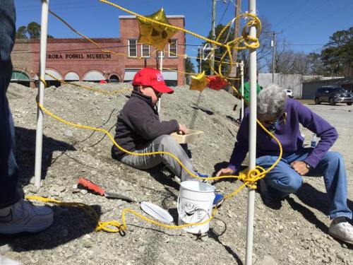 A child in a red hat sits on gravel, sifting through dirt, while an adult nearby examines the ground at a fossil museum site.