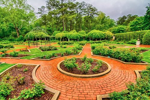 A beautifully landscaped garden with brick pathways, lush greenery, and neatly arranged flower beds under a cloudy sky.