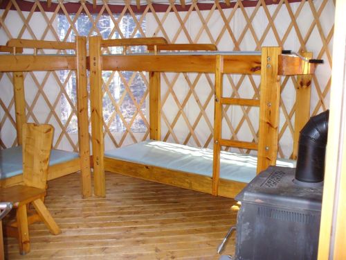 Interior of a yurt featuring wooden bunk beds, a small table, and a stove on a wooden floor. Natural light from windows.