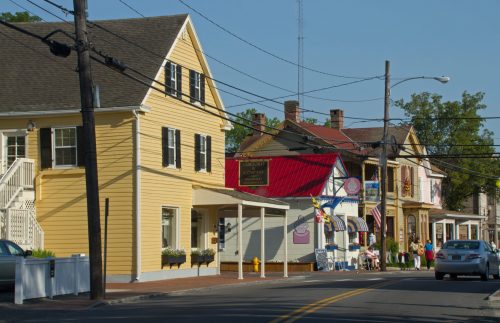 A quaint street scene featuring yellow houses, shops, and people enjoying a sunny day. Power lines crisscross overhead.