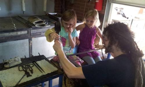 A person with dreadlocks demonstrates glassblowing to two young girls in a workshop setting.