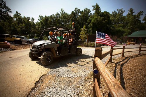 A group of people in a UTV driving on a gravel road, with an American flag and trees in the background.
