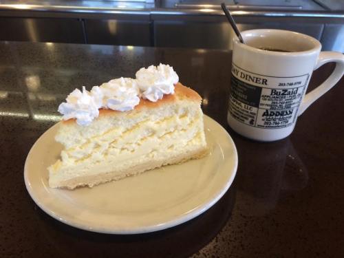 A slice of creamy cheesecake topped with whipped cream, served with a cup of coffee on a diner table.