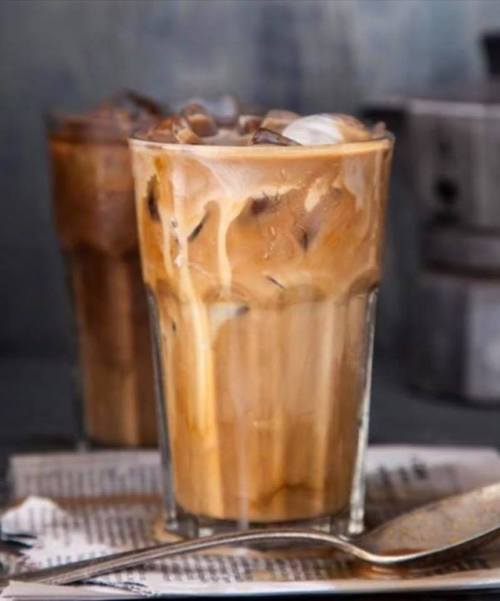 A tall glass of iced coffee with cream swirling, set on a table with a spoon and newspaper in the background.