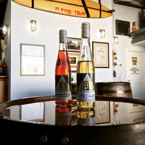 Two bottles of flavored beverages, one red and one yellow, displayed on a wooden barrel in a cozy, decorated space.