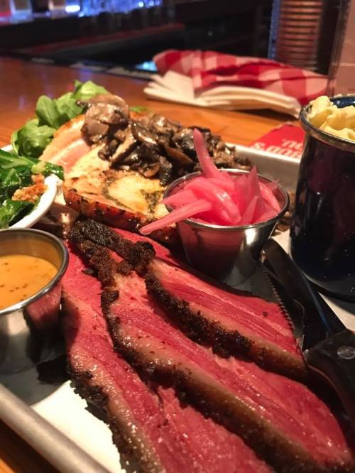 A platter featuring sliced smoked meat, pickled onions, sautéed mushrooms, greens, and a side of sauce.