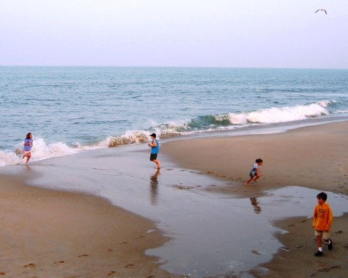 A beach scene with four children playing near the water, waves gently lapping at the shore under a clear sky.