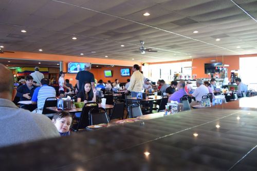A busy restaurant scene with diners at tables, bright orange walls, and TVs showing sports in the background.