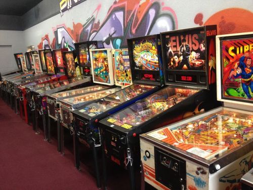 A row of vintage pinball machines with colorful designs and graffiti on the wall behind them.
