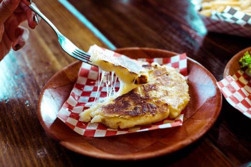 A fork lifting a cheesy, golden-brown dish from a wooden plate lined with red and white checkered paper.
