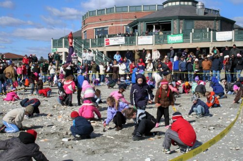 A crowded beach scene with children digging in the sand, wearing colorful winter clothing, and a building in the background.