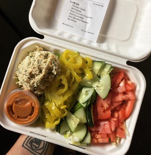 A takeout container with chicken salad, cucumbers, tomatoes, yellow peppers, and a dressing cup.