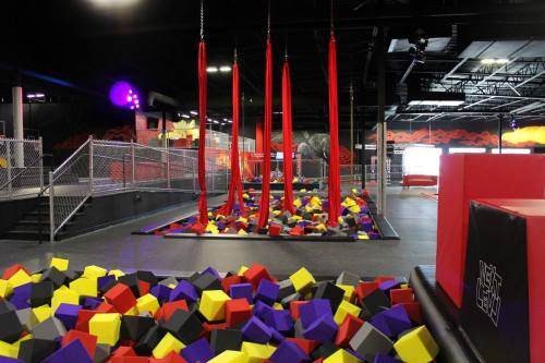 A vibrant indoor trampoline park featuring colorful foam pits and hanging red aerial silks.