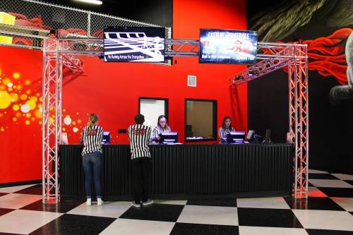 Two staff members in striped shirts work at a reception desk in a vibrant indoor facility with screens displaying information.