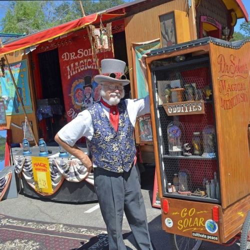 A magician in a top hat and vest stands beside a colorful trailer displaying oddities and magic props.