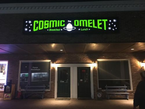 Exterior of "Cosmic Omelet" restaurant, featuring a bright green sign and a brick facade, open for breakfast and lunch.