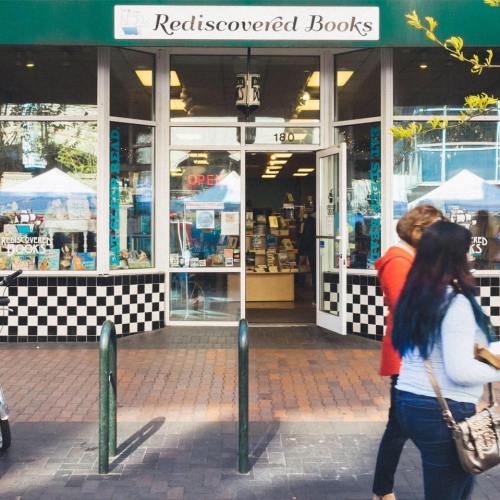 A bookstore entrance with a green awning, people walking by, and a checkered sidewalk. Signs indicate it's open.