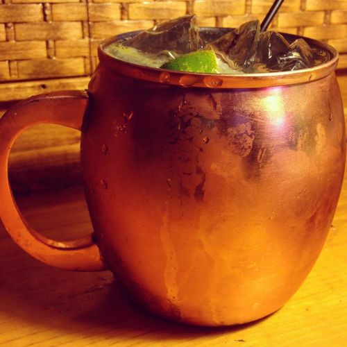 A copper mug filled with ice and a lime wedge, resting on a wooden surface with a woven background.