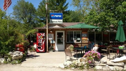 A rustic store with a Pepsi sign, outdoor seating, and a vending machine, surrounded by greenery.