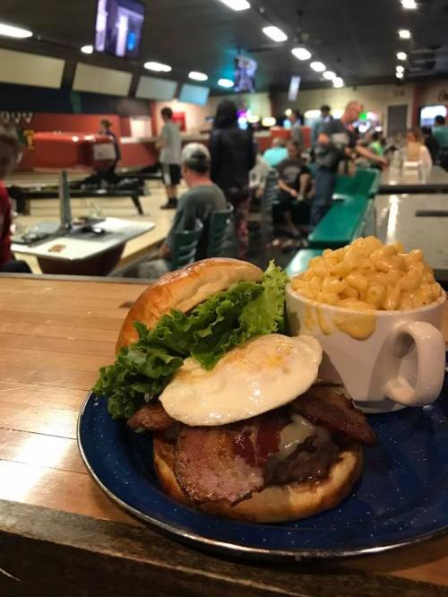 A delicious burger with bacon, lettuce, and a fried egg, served with a side of macaroni and cheese in a bowling alley.