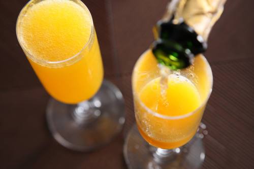 Two glasses of orange mimosa with bubbles, one being poured from a bottle.