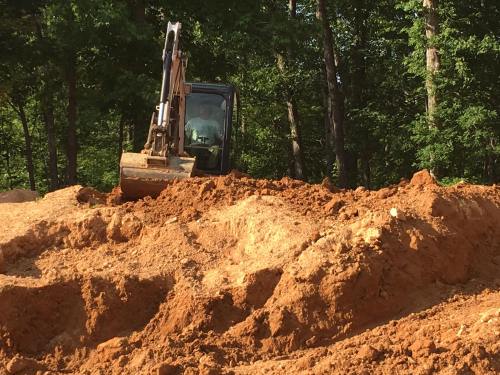 Excavator working on a dirt mound in a wooded area, surrounded by trees.