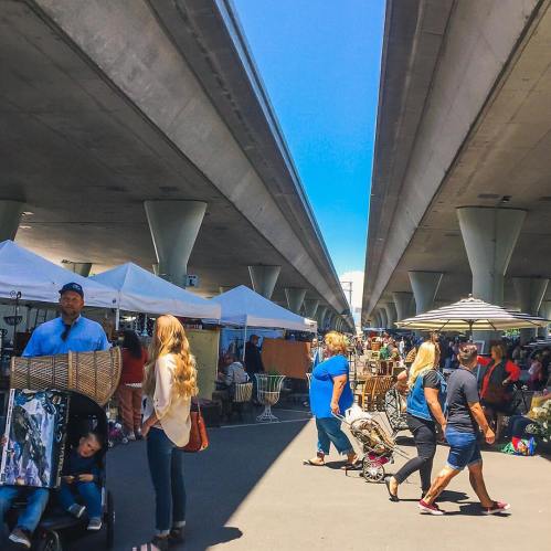 A bustling market scene under a highway, with vendors, shoppers, and tents lining the walkway on a sunny day.
