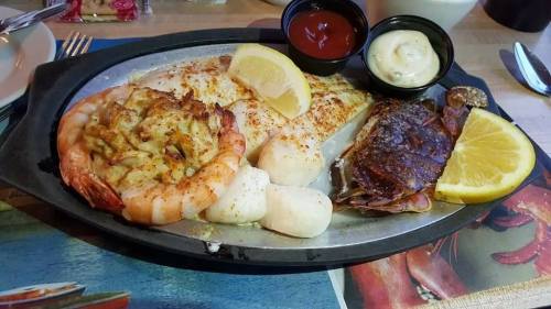 A sizzling seafood platter with shrimp, fish, crab, and lemon slices, served with dipping sauces on a metal tray.