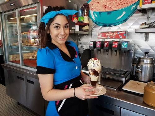 A smiling server in a blue uniform holds a sundae with chocolate sauce and whipped cream at an ice cream shop.