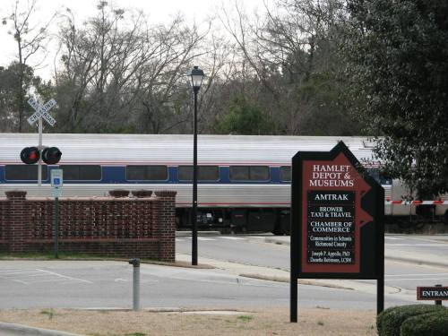 A train passes by a sign for Hamlet Depot & Museums, with information about Amtrak and local commerce.