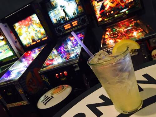 A refreshing cocktail with a lemon slice sits on a table in front of colorful pinball machines.