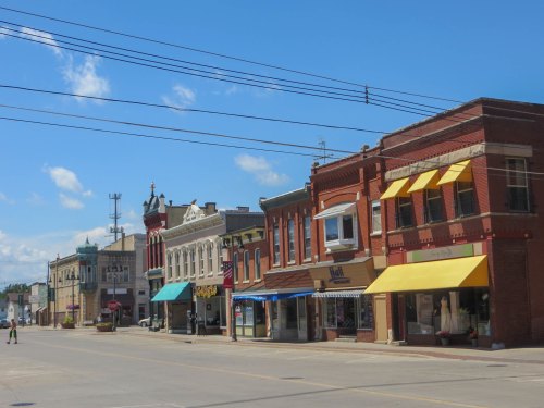 A sunny street view of a small town with historic brick buildings and storefronts under a blue sky.