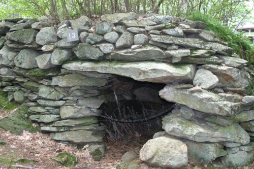 A stone structure with a small entrance, surrounded by greenery and numbered 13, resembling a rustic shelter or storage.