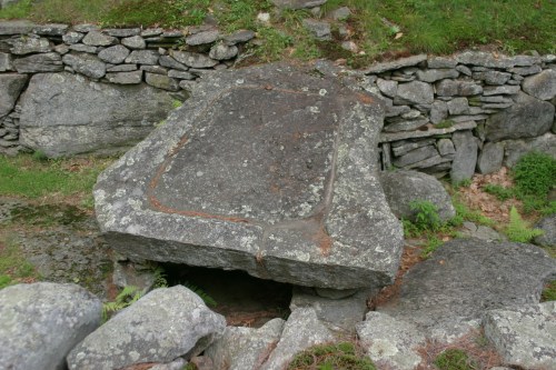 A large, flat stone resting on smaller rocks, surrounded by a low stone wall and greenery.