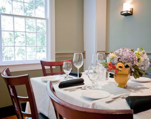 A dining table set with white tablecloth, glassware, and a floral centerpiece, near a window with natural light.