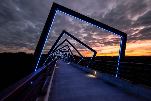 A modern bridge with illuminated geometric frames, set against a dramatic sunset and cloudy sky.