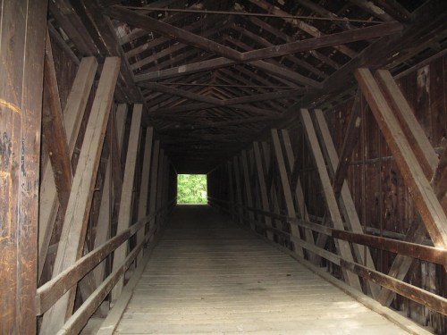 A view inside a wooden covered bridge, showcasing beams and a lighted exit at the far end.