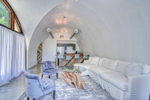 A modern, curved interior featuring a white sofa, glass coffee table, and stylish chairs, with large windows and a kitchen area.