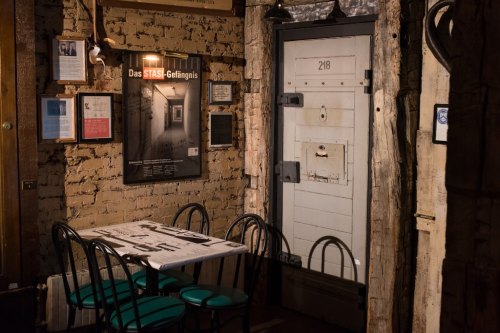 A dimly lit room featuring a table with chairs and a door labeled "218," showcasing historical STASI prison artifacts.