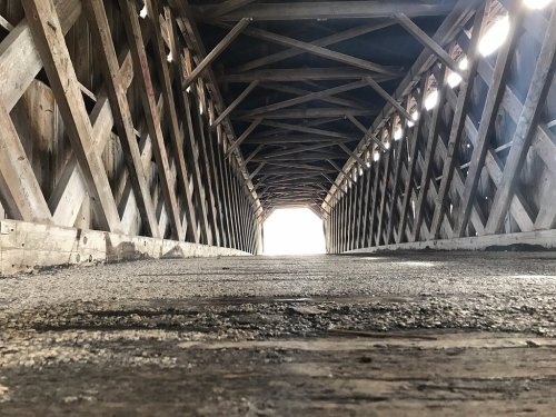 View from inside a covered bridge, showcasing wooden beams and a light-filled opening at the far end.