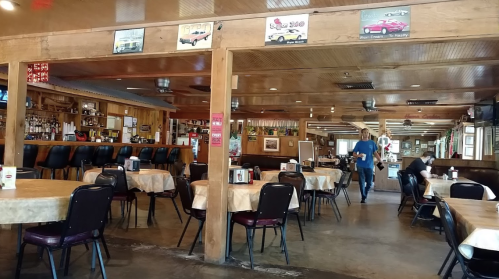 Interior of a casual restaurant with wooden decor, tables set for dining, and a bar area in the background.