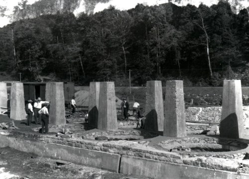 Workers constructing stone structures in a forested area, with machinery and tools visible in the background.