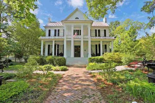 A grand white mansion with a porch, surrounded by lush greenery and a brick pathway under a blue sky.