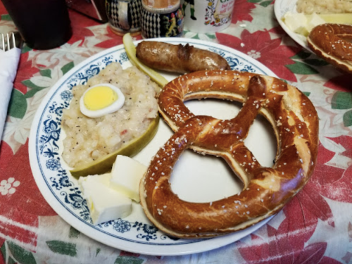 A plate with a large pretzel, sausage, potato salad with an egg, and cheese, set on a floral tablecloth.