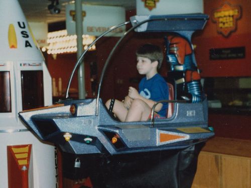 A young boy sits in a futuristic arcade ride, surrounded by colorful decorations and a rocket display.