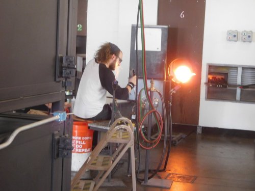 A person sits on a ladder, using a torch near a glowing furnace in a workshop. Tools and equipment are visible around them.