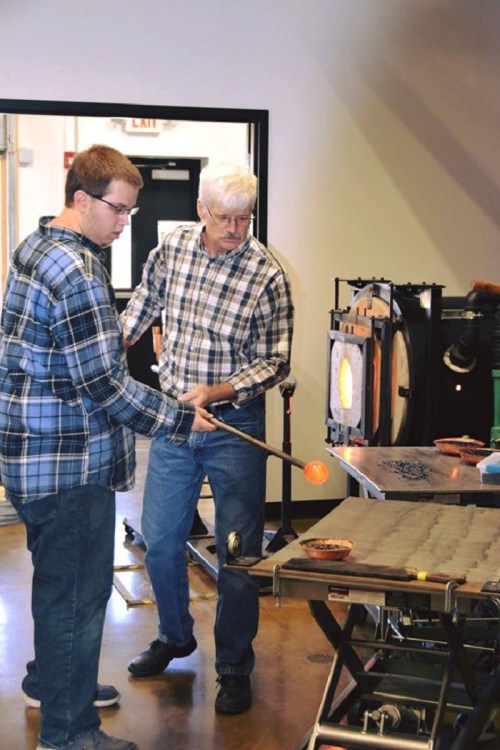Two men work together in a glassblowing studio, shaping molten glass with a blowpipe near a furnace.