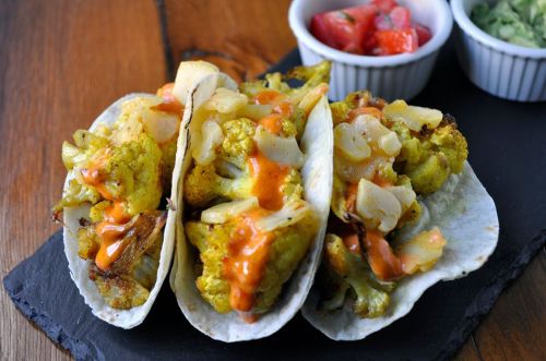 Three cauliflower tacos topped with onions and sauce, served on a slate plate with bowls of salsa and guacamole.