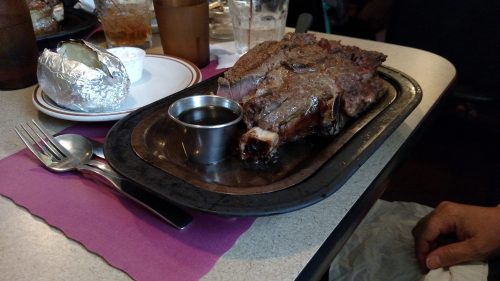 A sizzling steak on a metal platter with a small cup of sauce, accompanied by a baked potato in foil.