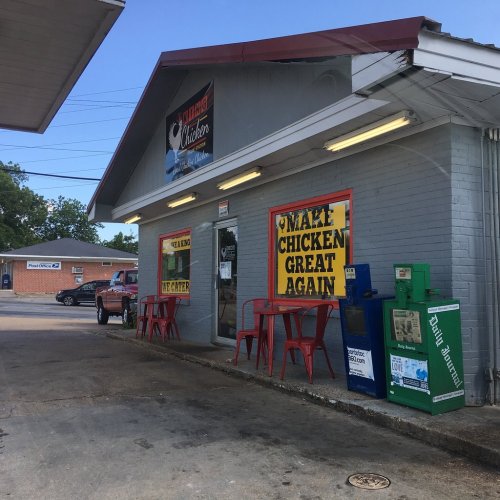 A small chicken restaurant with colorful signage, outdoor seating, and newspaper stands in front.